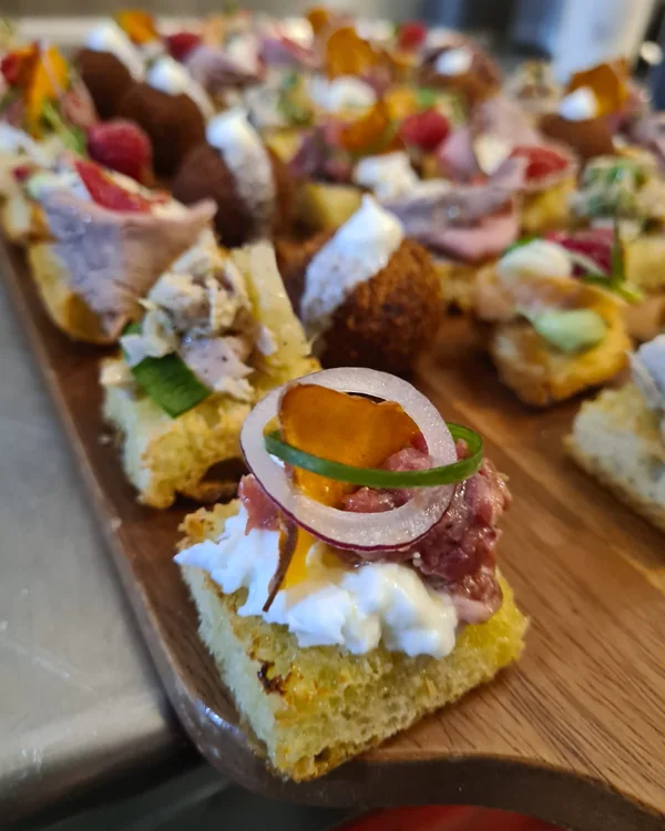 Close-up of a square of toasted bread with stracciatella, raw beef, a ring of red onion and a sweet potato crisp, more canapés behind
