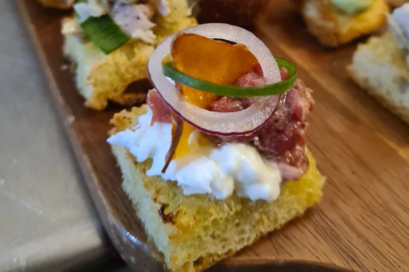 Close-up of a square of toasted bread with stracciatella, raw beef, a ring of red onion and a sweet potato crisp, more canapés behind
