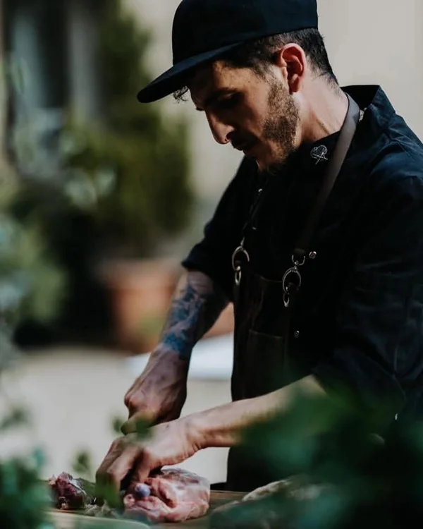 Marco Genna cutting meat on a board outdoors, herbs out of focus in the foreground