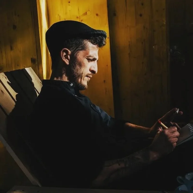 Marco Genna seated against a wooden wall in warm light, writing in a notebook