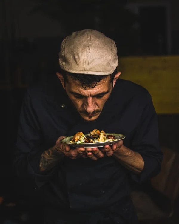 Marco Genna in a dark kitchen, holding a plate close and smelling it before service