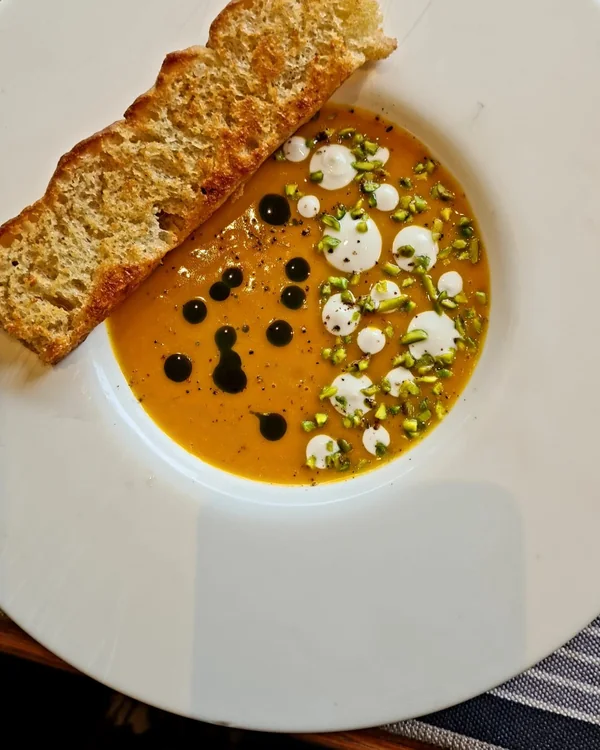 Orange pumpkin soup in a wide white bowl, dotted with cream, dark oil and chopped pistachio, with a slice of toasted bread