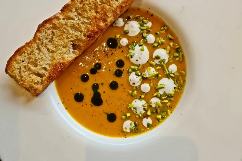 Orange pumpkin soup in a wide white bowl, dotted with cream, dark oil and chopped pistachio, with a slice of toasted bread