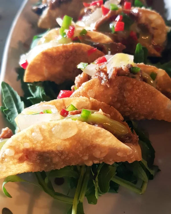 A row of small fried taco shells filled with shredded meat, onion and diced peppers, on rocket