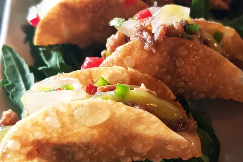 A row of small fried taco shells filled with shredded meat, onion and diced peppers, on rocket