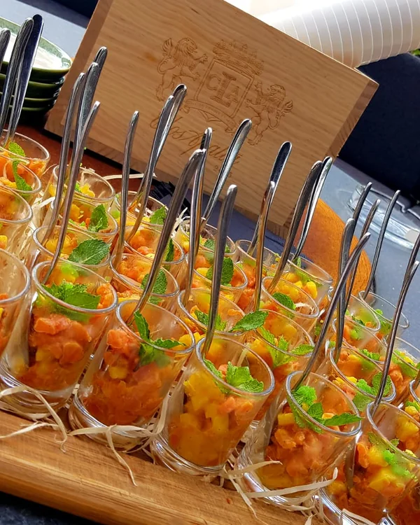Rows of small glasses of salmon tartare with yellow fruit and mint, each with a spoon, on a wooden board outdoors