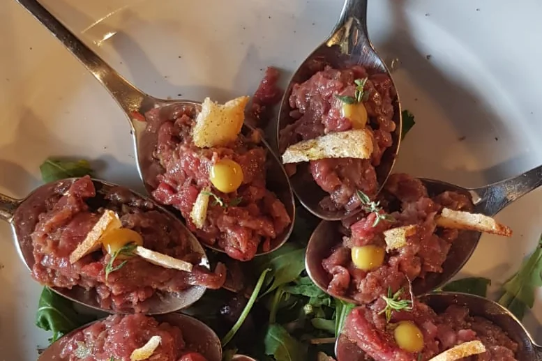 Eight spoons of beef tartare arranged in a circle on a white plate, each with a dot of yellow sauce, thyme and a thin crisp